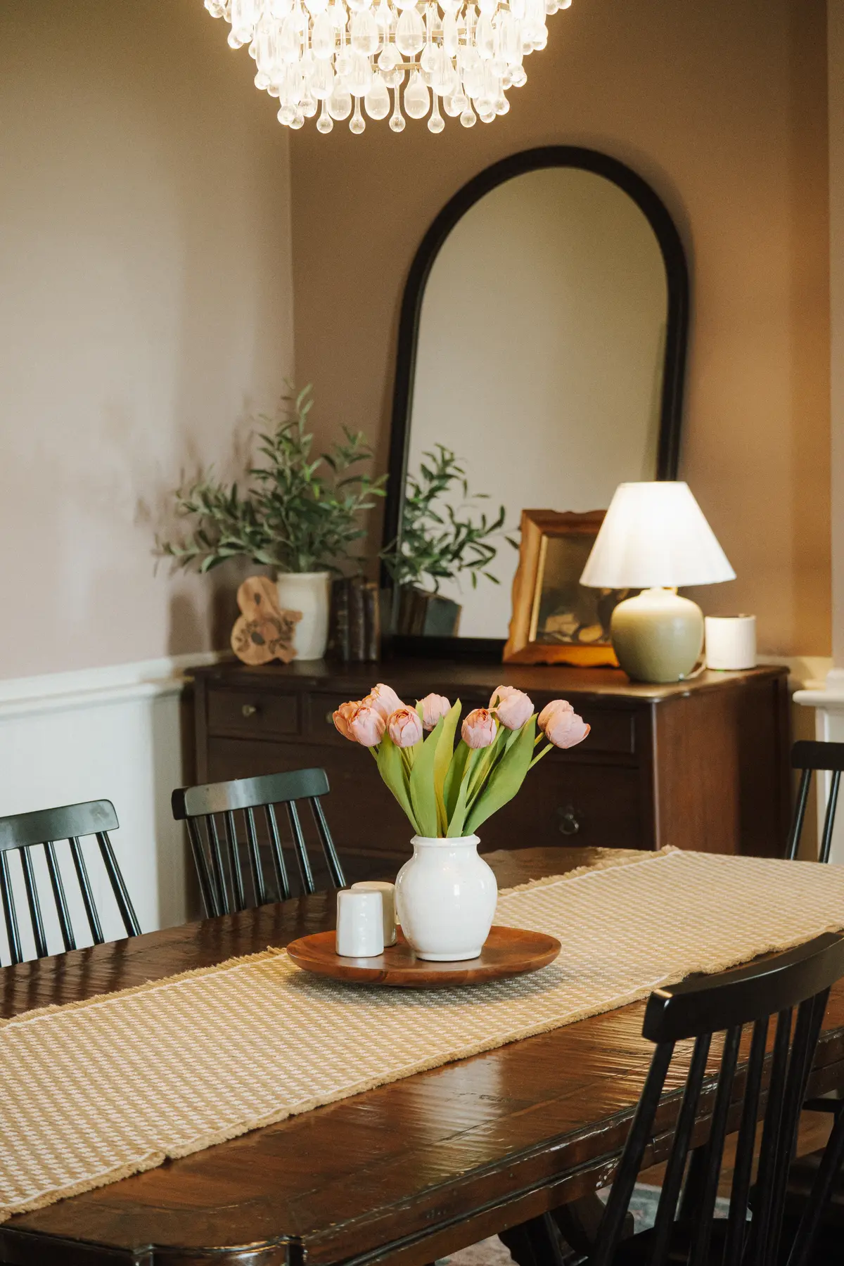 Dining table set with pink tulips beneath an arched black mirror and a ceramic table lamp
