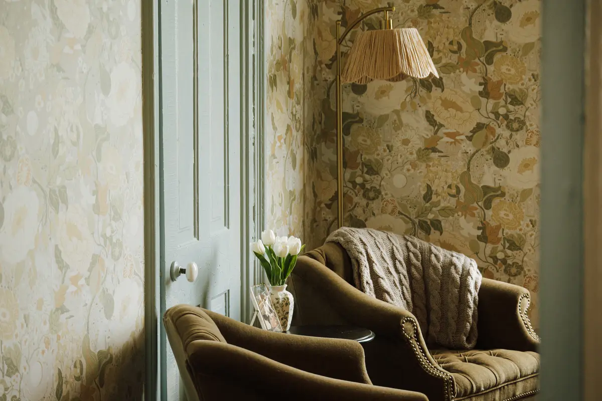 Botanical wallpapered corner with velvet armchairs, a pleated floor lamp, and white tulips