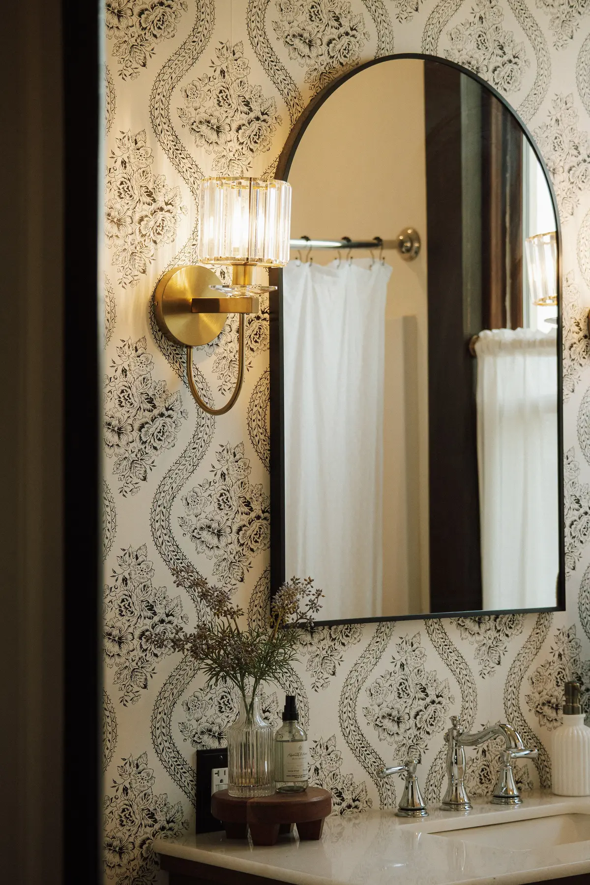 Powder bath with patterned wallpaper, an arched black mirror, and a crystal brass sconce